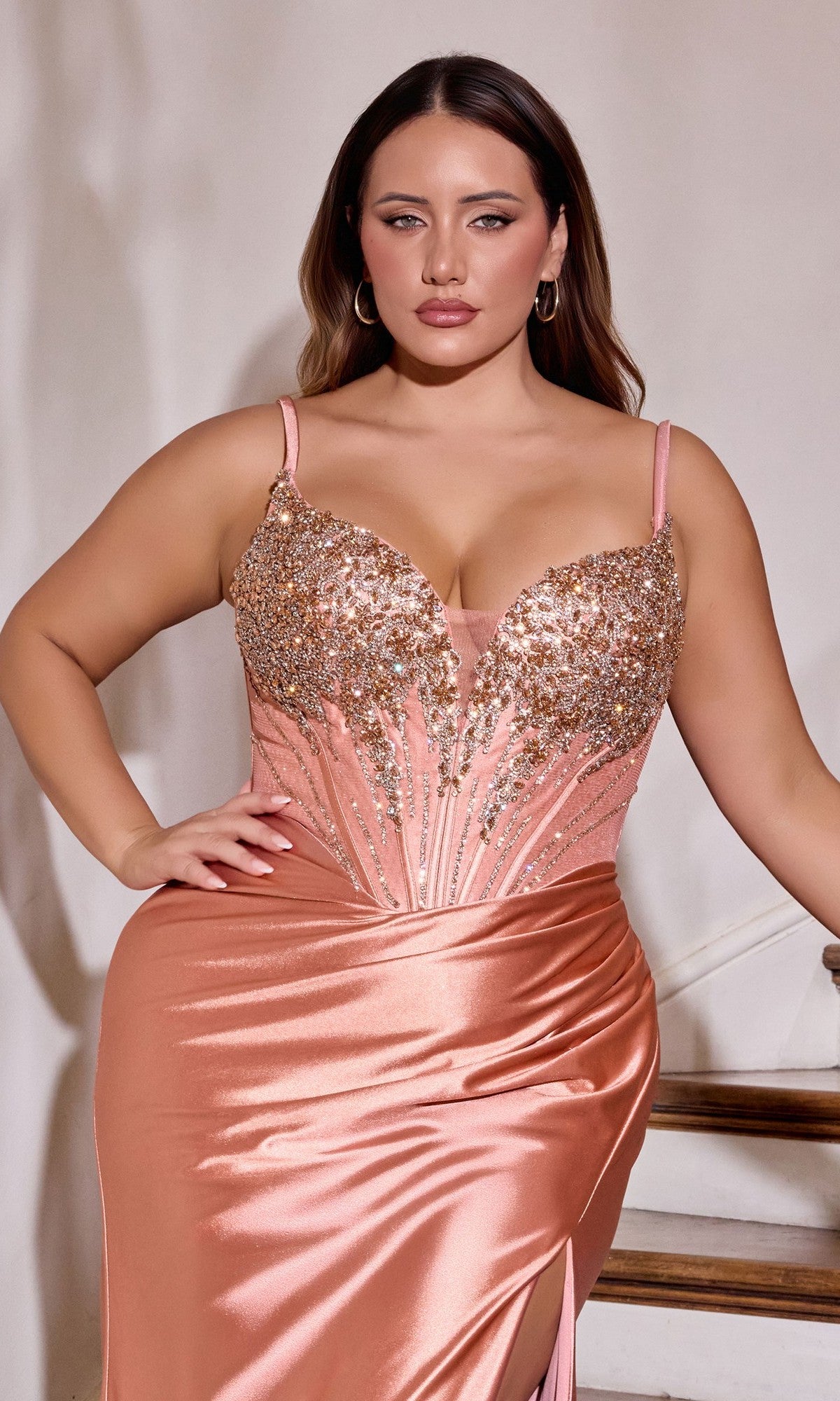 A woman wearing the Long Plus-Size Prom Dress: Ladivine CD729C in peach stretch satin with a beaded corset bodice and gold sequin embellishments stands with one hand on her hip.